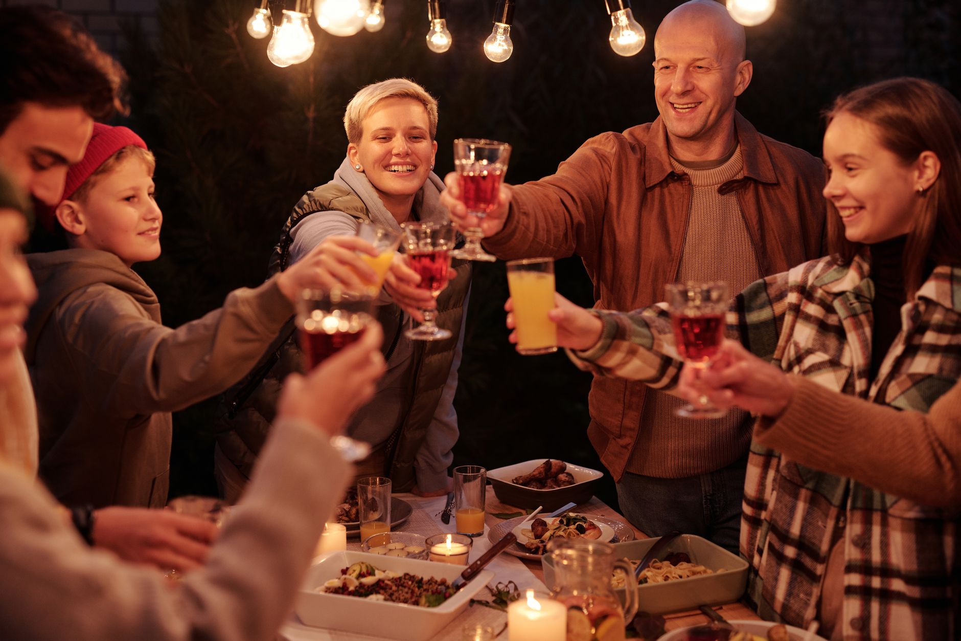 Three generations of a family raising glasses together under warm string lights in a backyard.